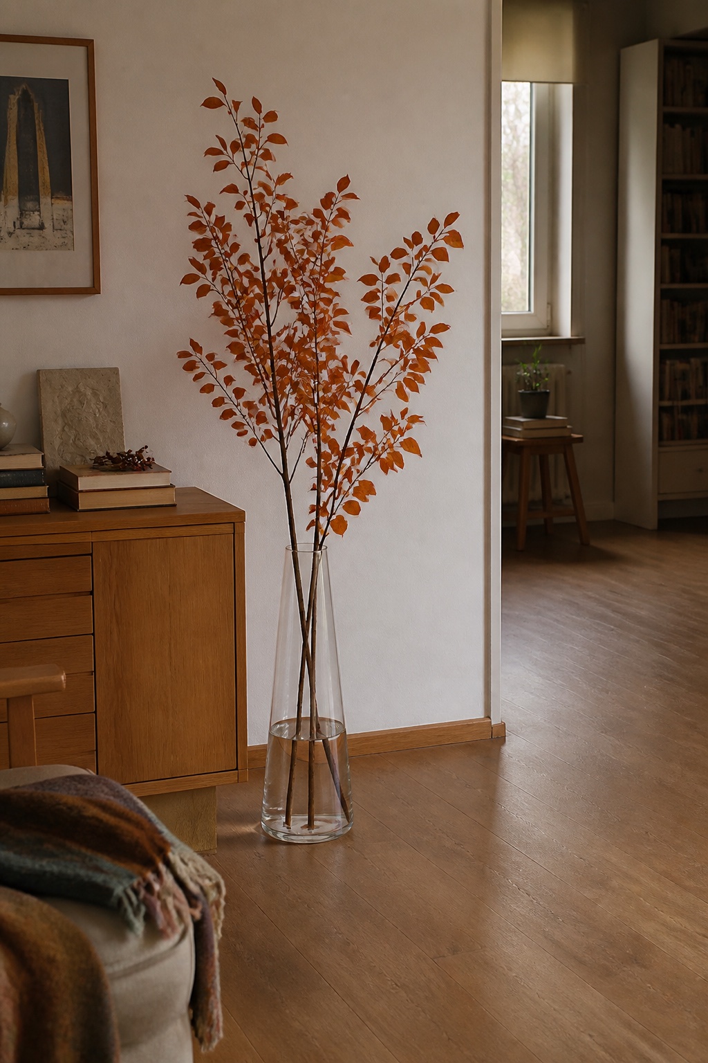 Hohe orangefarbene Herbstzweige in einer klaren Bodenvase neben einem Sideboard