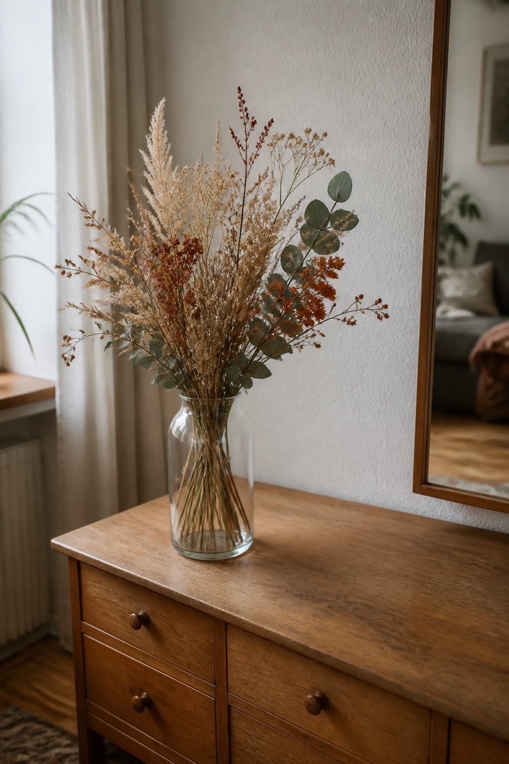 Herbstlicher Trockenblumenstrauß mit Eukalyptus in einer klaren Glasvase auf einer Kommode