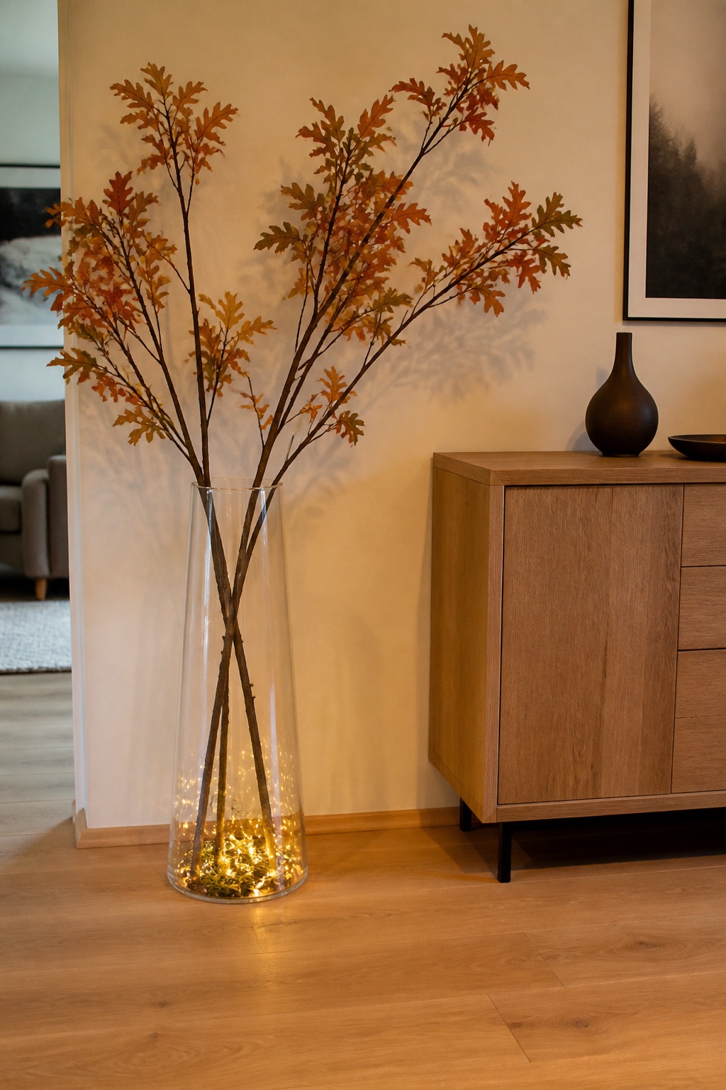 Hohe klare Bodenvase mit Herbstzweigen und Licht am Glasboden neben einem Sideboard