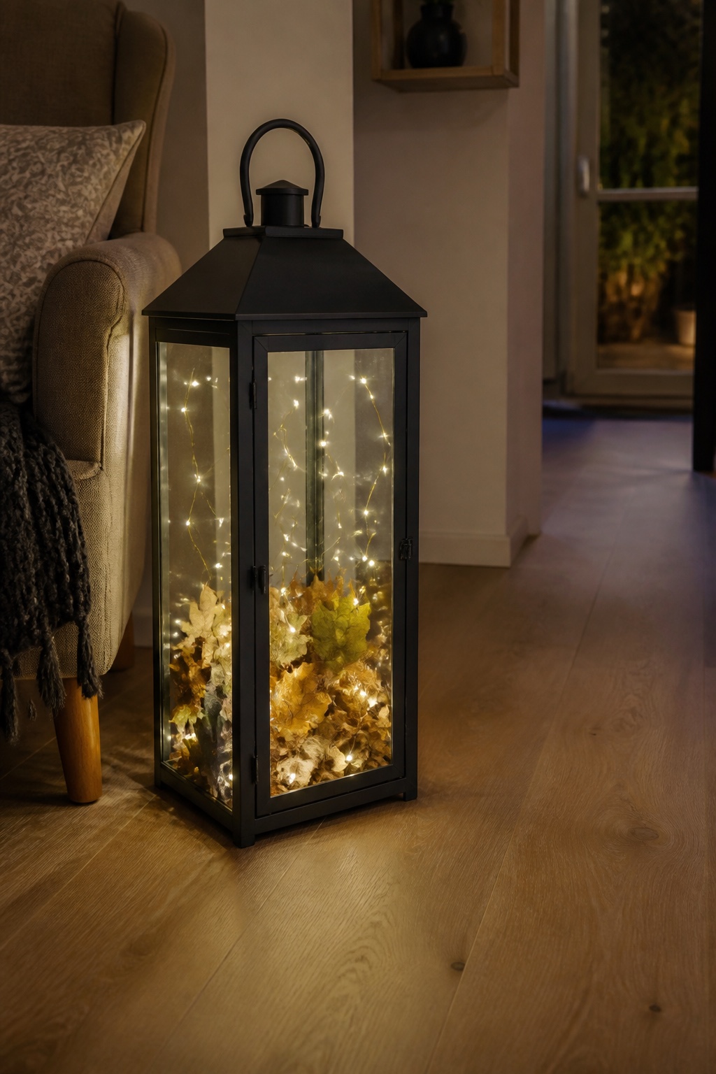 Glaslaterne mit warmweißer Lichterkette und trockenen Blättern neben einem Sessel