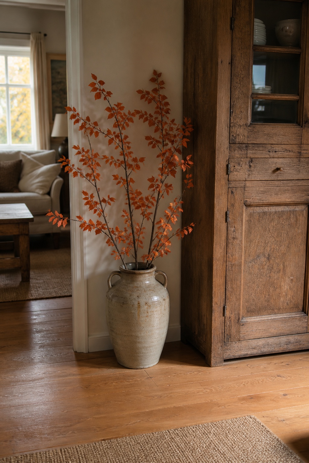 Hohe orange Herbstzweige in einem schweren cremefarbenen Keramikkrug neben einem Holzschrank